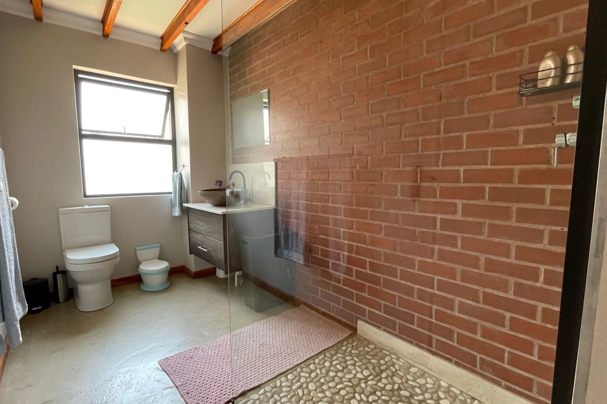 Modern bathroom featuring an exposed brick feature wall, glass-enclosed shower with pebble flooring, floating vanity with vessel sink, and exposed timber ceiling beams.