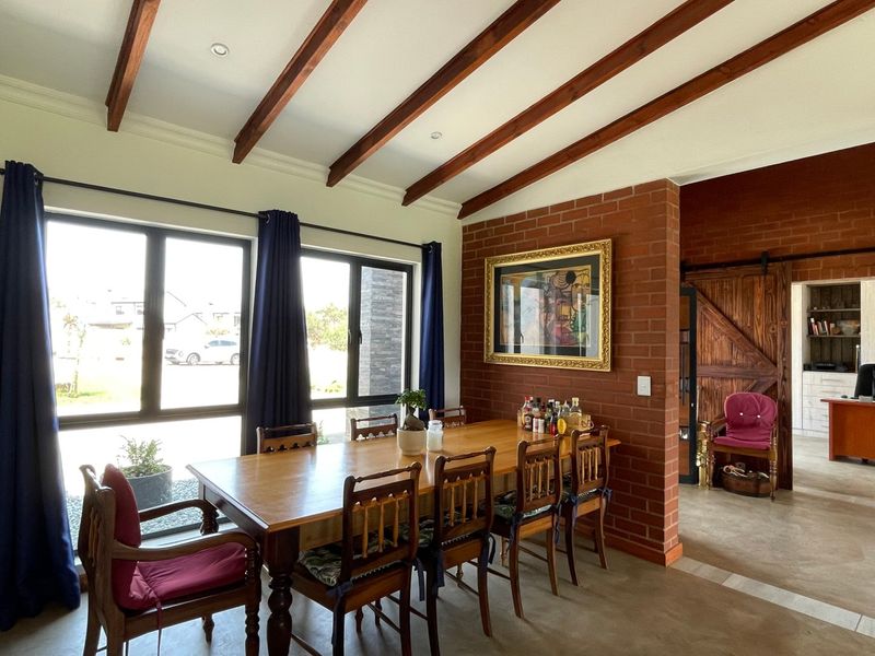 Spacious dining area featuring exposed timber ceiling beams, a rustic brick feature wall, polished concrete floors, and large windows providing natural light.