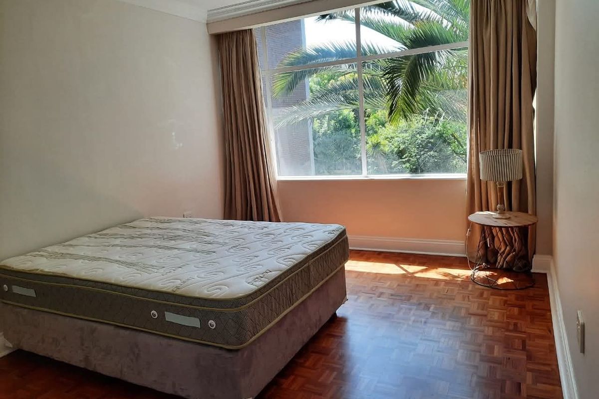 A bright bedroom featuring classic parquet flooring, large windows with natural light, and views of surrounding greenery.