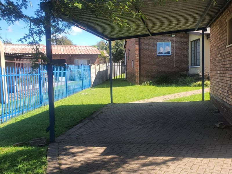 A covered carport area with paved flooring, situated alongside a brick residential building and enclosed by metal fencing.
