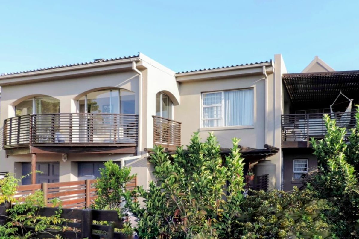 Rear elevation of a multi-story house featuring timber balconies, arched window detailing, and a pergola-covered outdoor area set within a landscaped garden.