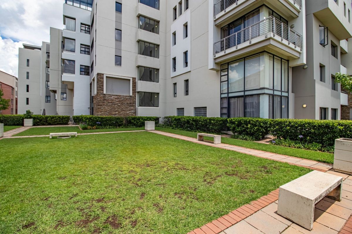 A well-maintained communal garden area featuring paved walkways and manicured lawns, situated between modern apartment building facades with stone accents and balconies.