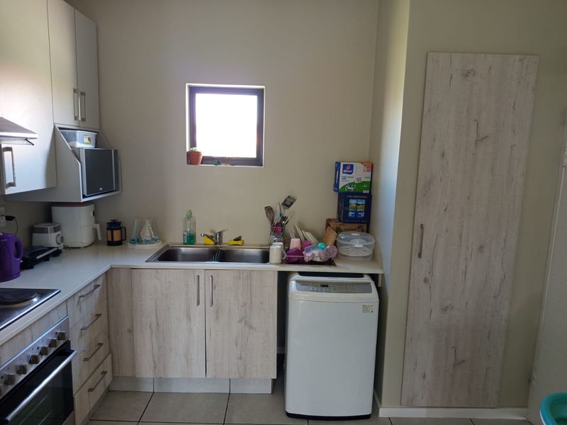 A functional kitchen featuring light wood-grain cabinetry, built-in oven and hob, stainless steel sink, and a tall pantry cupboard with tiled flooring.