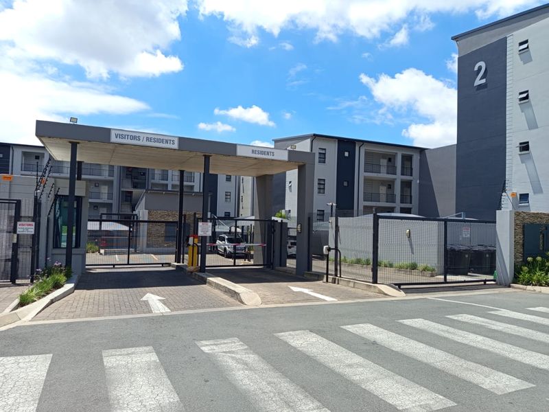 The main entrance to the apartment complex featuring a secure gated access point, paved driveway, and modern multi-story residential building facade.
