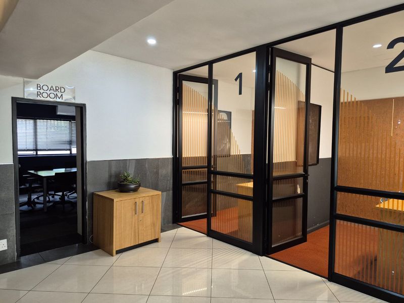 Modern office hallway featuring glass-partitioned meeting rooms with black frames and tiled flooring, providing a professional and organized workspace environment.