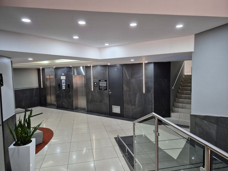 Modern apartment building lobby featuring tiled flooring, stainless steel elevator doors, integrated wall lighting, and a staircase with glass balustrades.