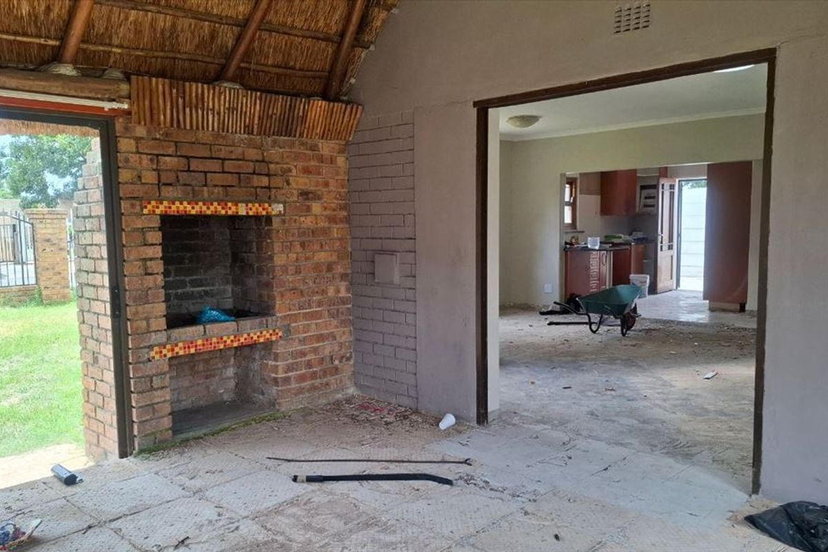 An open-plan living area featuring a built-in brick fireplace and thatched roof, currently undergoing renovation with unfinished flooring and direct access to the garden.