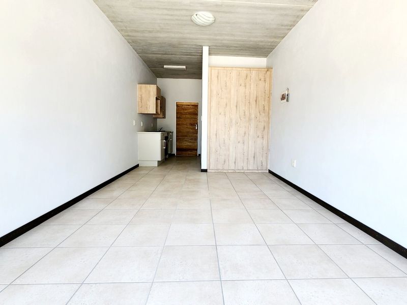 An open-plan studio apartment featuring tiled flooring, exposed concrete ceilings, and built-in cabinetry with a compact kitchen area.