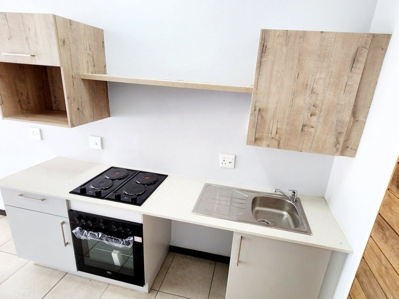 A modern, compact kitchen featuring light wood-finish cabinetry, stone countertops, a built-in electric oven and hob, and a stainless steel sink.