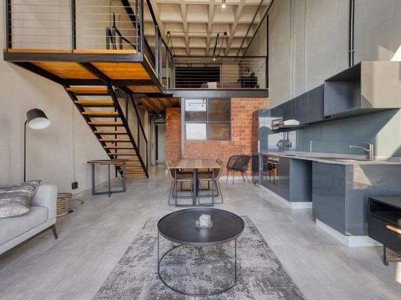 Modern industrial open-plan living area featuring a mezzanine level, exposed brickwork, concrete floors, and a sleek contemporary kitchen with built-in cabinetry.