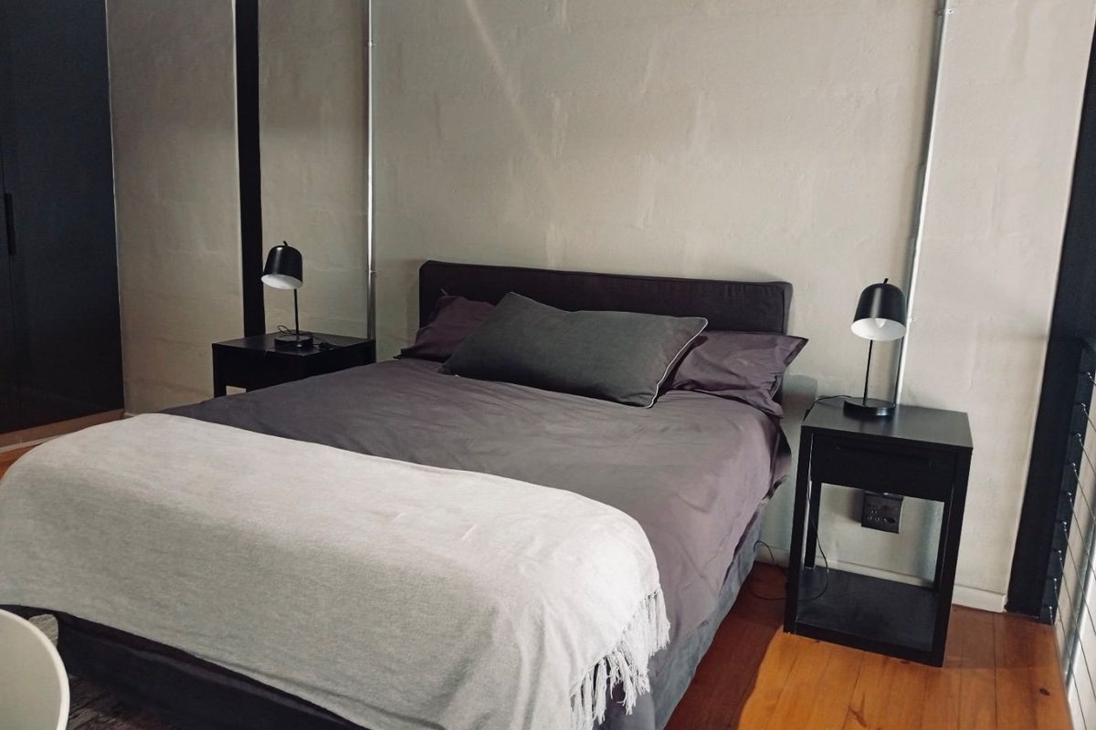 A modern bedroom featuring industrial-style exposed electrical conduit, warm timber flooring, and a neutral painted block wall finish.