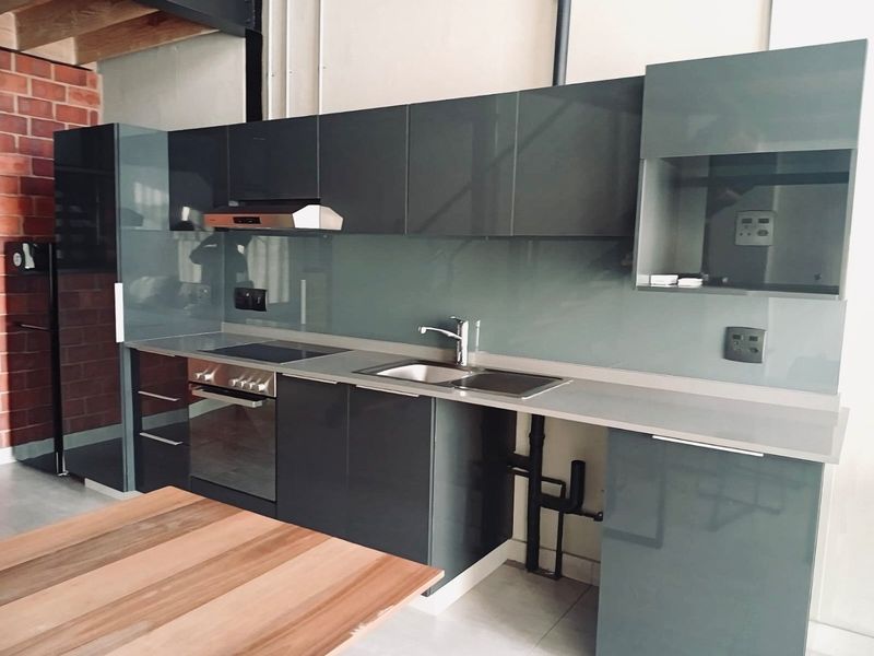 Modern kitchen featuring dark grey high-gloss cabinetry, glass splashback, integrated appliances, and an industrial aesthetic with exposed brick and structural beams.