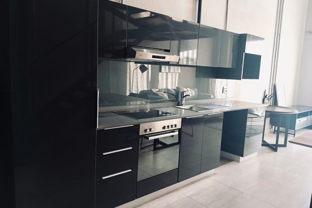 Modern kitchen featuring sleek glossy black cabinetry, integrated oven, electric hob, extractor fan, and a reflective mirror splashback with stone countertops.