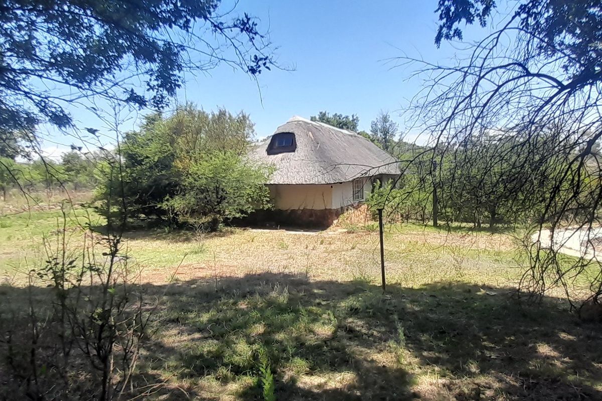 A view of a rustic cottage with a traditional thatch roof and stone base, situated within a natural, overgrown farm landscape.