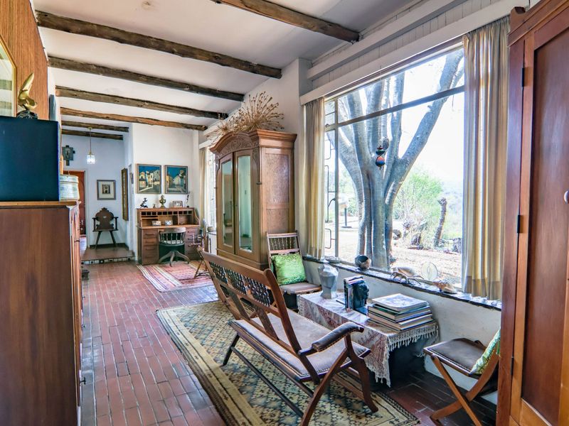 A rustic living area featuring exposed timber ceiling beams, terracotta tile flooring, and a large picture window overlooking the farm landscape.