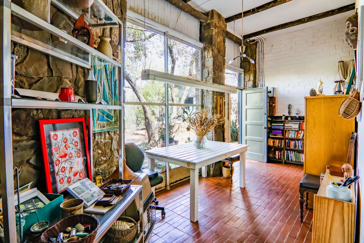 A rustic living space featuring exposed stone walls, timber ceiling beams, terracotta flooring, and large windows providing natural light and garden views.