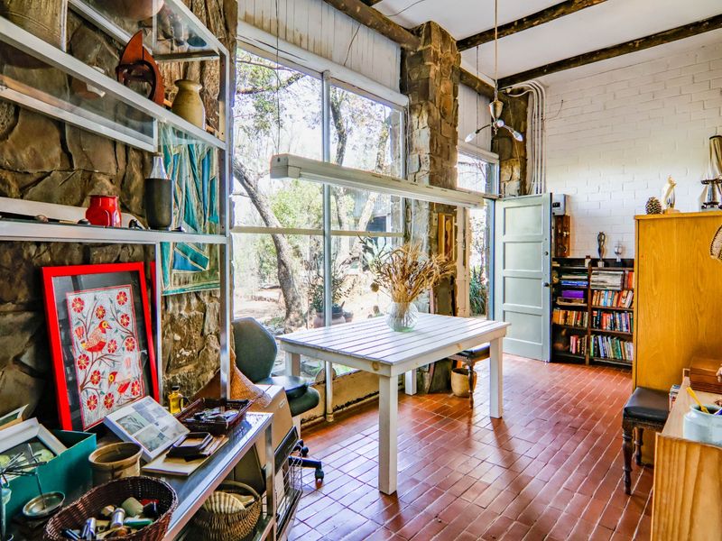 A rustic living space featuring exposed stone walls, timber ceiling beams, terracotta flooring, and large windows providing natural light and garden views.