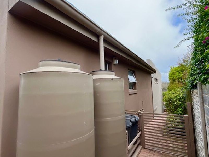 Side exterior area featuring two large water storage tanks connected to the roof guttering system, with paved flooring and a slatted gate.