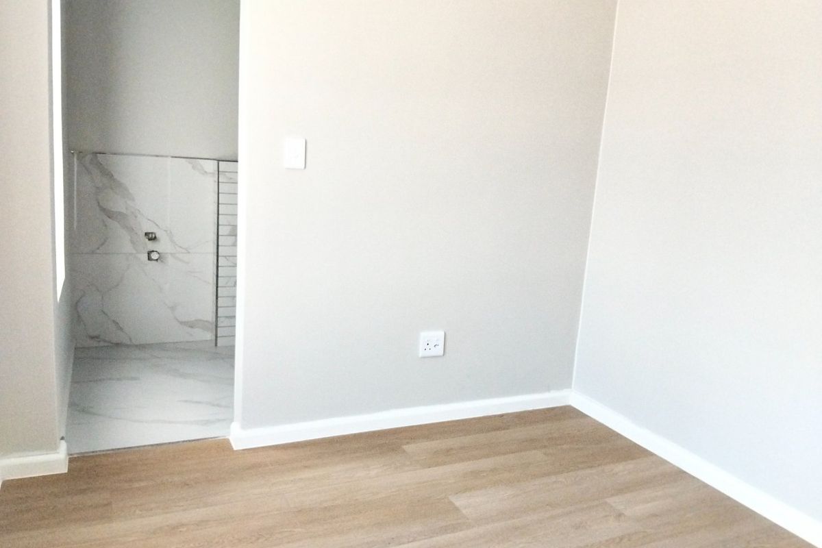A bright, empty bedroom featuring light wood-look flooring and a doorway leading into an en-suite bathroom with marble-style wall tiling.