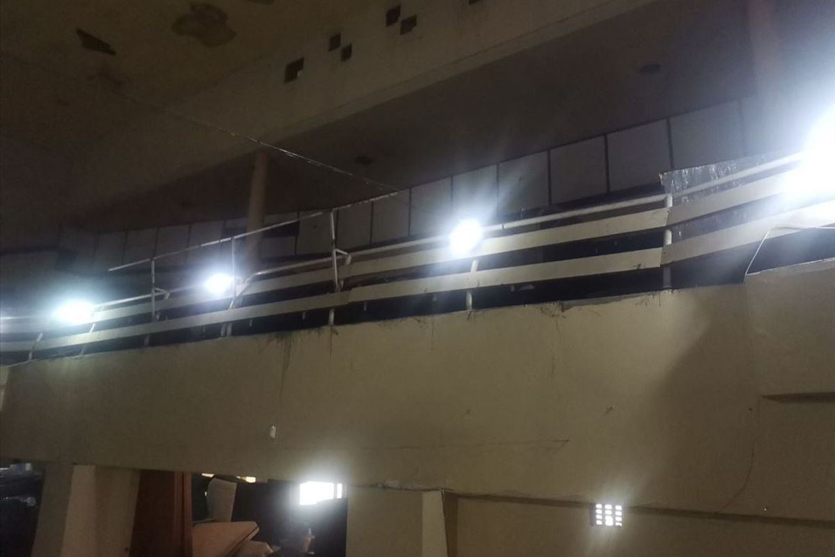 A dimly lit, dilapidated interior space featuring a mezzanine level with railings and significant signs of structural wear and ceiling damage.
