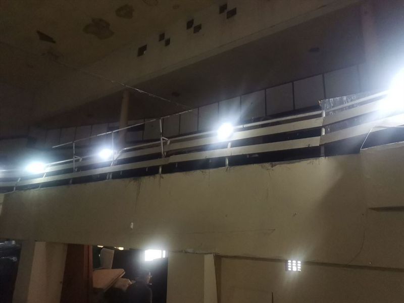 A dimly lit, dilapidated interior space featuring a mezzanine level with railings and significant signs of structural wear and ceiling damage.