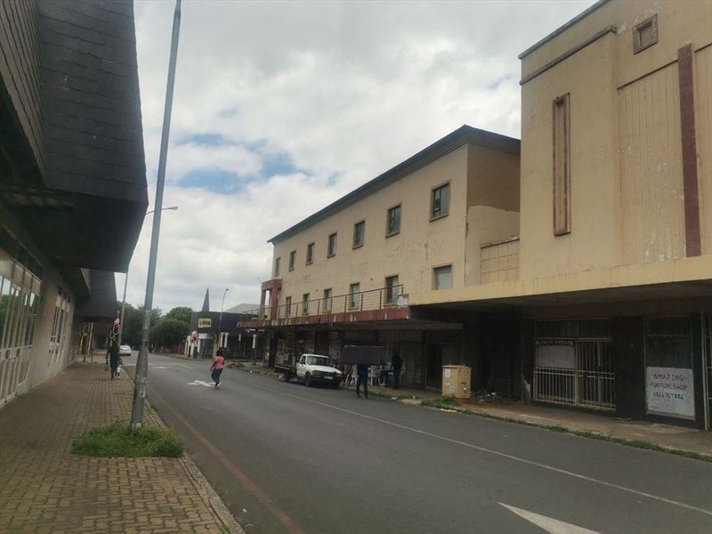 A street-level view of a commercial building facade with visible wear and tear, featuring a sidewalk and street infrastructure.