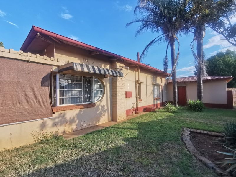 Rear view of the property featuring a brick facade, corrugated metal roofing, and established garden area with mature palm trees.