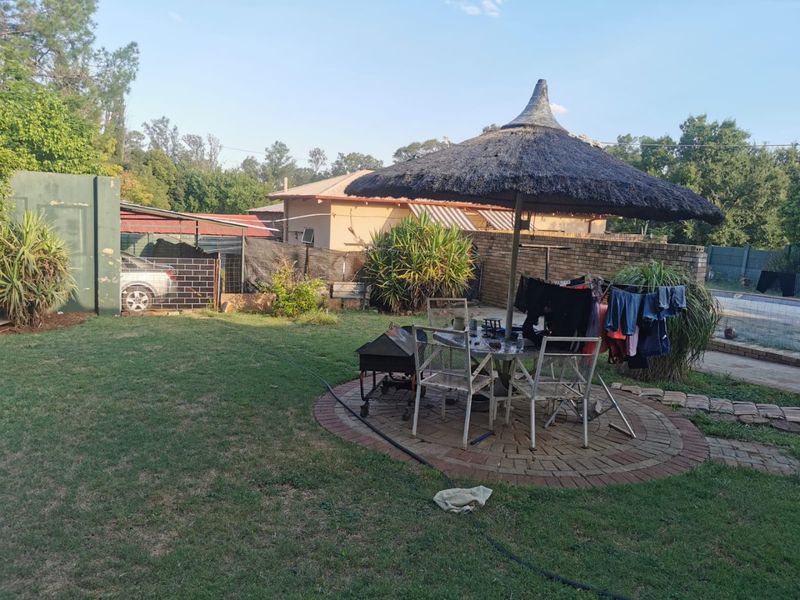 A private backyard garden featuring a circular brick-paved patio area with a thatched umbrella, enclosed by brick boundary walls and mature landscaping.