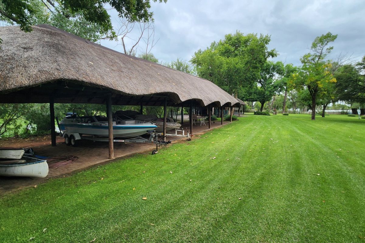 A spacious farm property featuring a long, covered thatch-roofed storage area for boats and equipment, set against a well-maintained expansive green lawn.