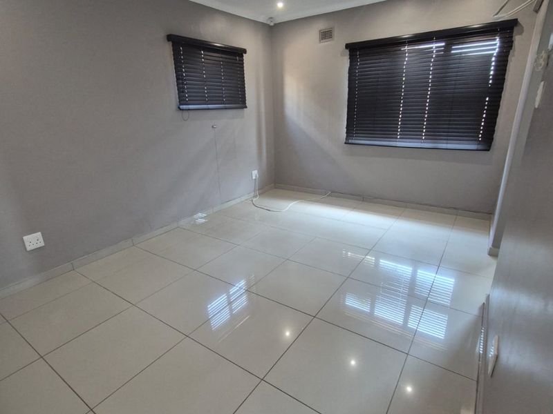 A bright, empty bedroom featuring neutral grey walls, polished porcelain floor tiles, recessed lighting, and fitted venetian blinds.
