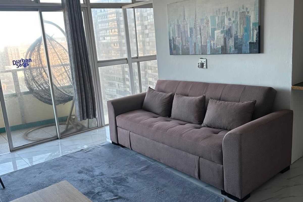 A bright living area featuring large floor-to-ceiling windows, tiled flooring, and a ceiling fan, offering natural light and access to the balcony.