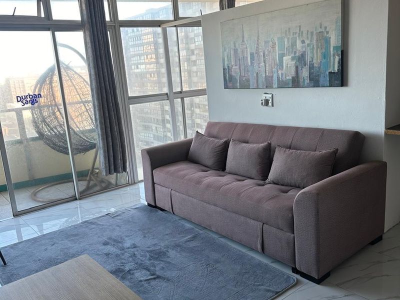 A bright living area featuring large floor-to-ceiling windows, tiled flooring, and a ceiling fan, offering natural light and access to the balcony.