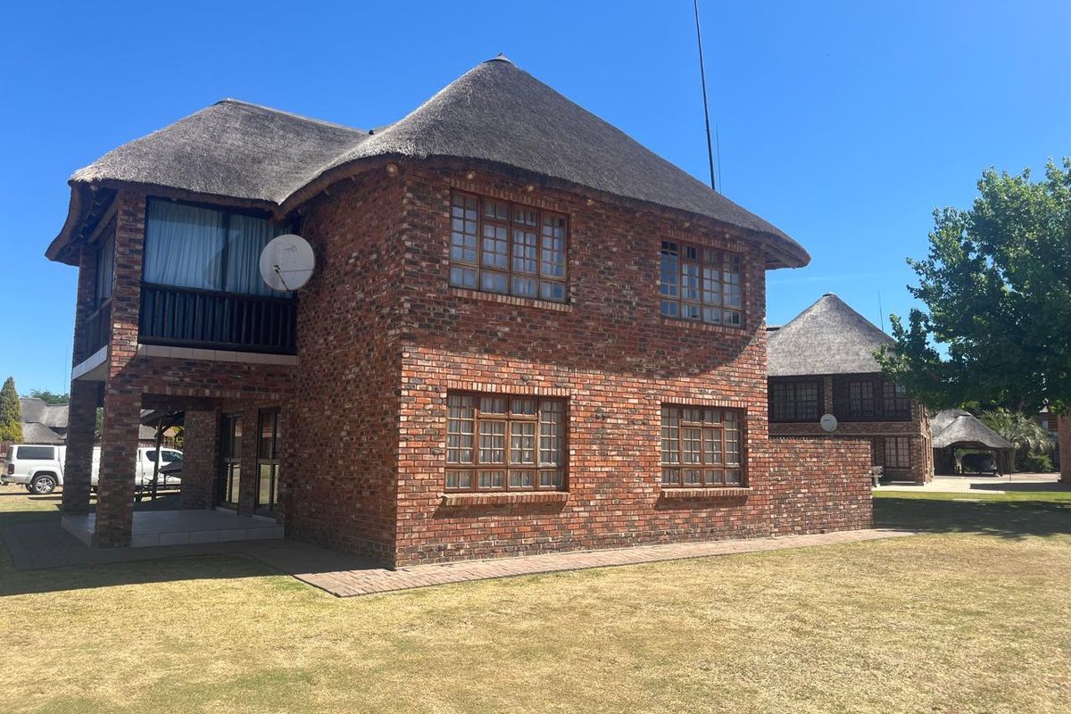 A two-story face brick house featuring a traditional thatch roof, timber-framed windows, and an upper-level balcony, set within a well-maintained garden area.