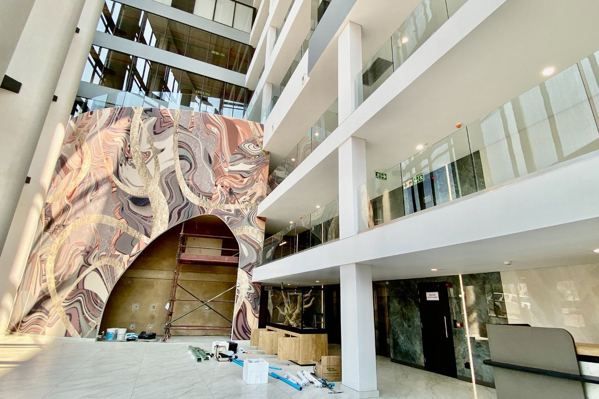 Spacious commercial atrium entrance featuring high ceilings, glass balustrades, marble-look flooring, and a striking decorative feature wall under construction.
