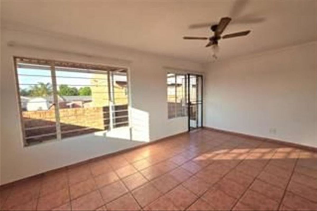 An empty living room featuring tiled flooring, a ceiling fan, and large windows with security bars, offering a bright and functional space.