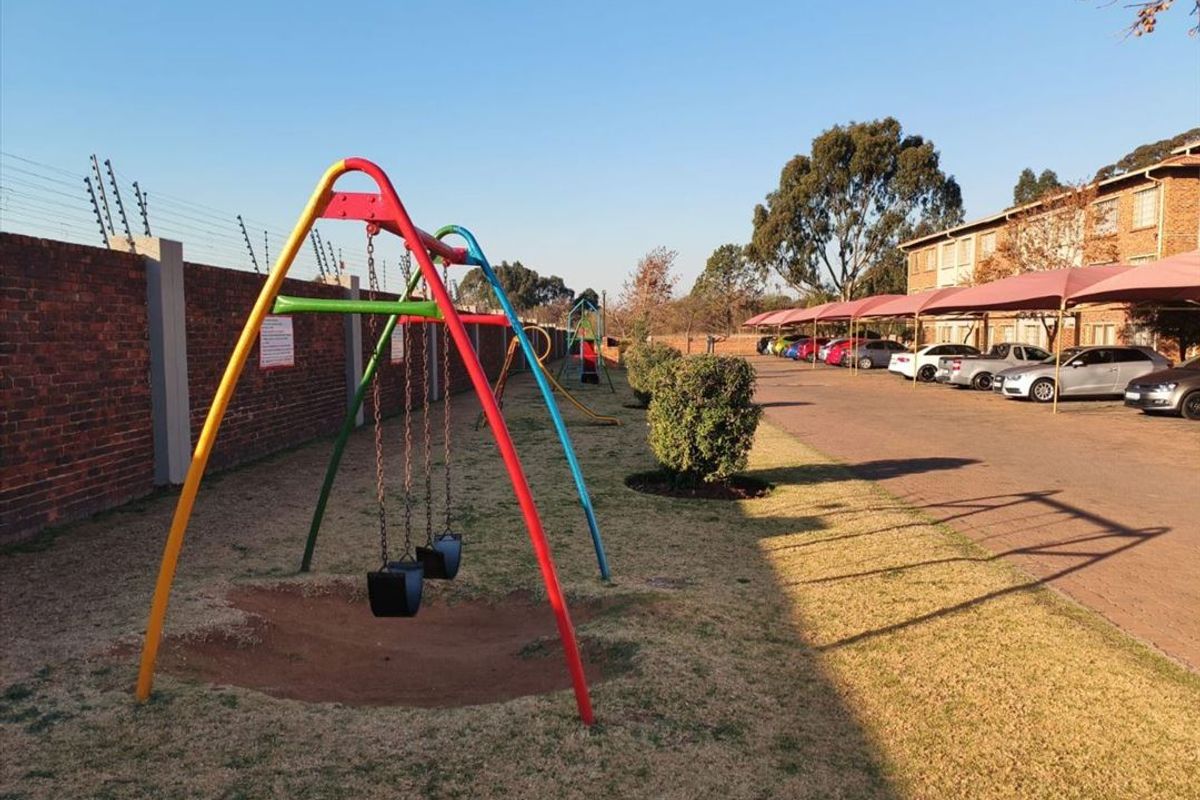 Exterior view of the apartment complex featuring a communal garden area with playground equipment, paved parking, and covered carports.