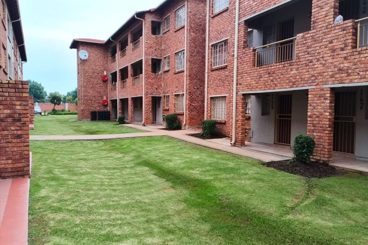 Exterior view of a multi-story face brick apartment complex featuring communal garden spaces, paved walkways, and private balconies for each unit.