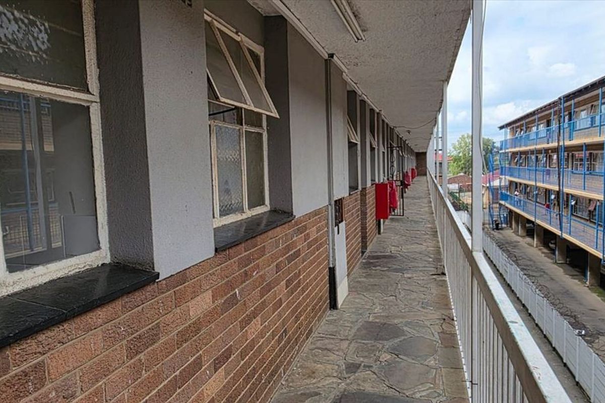 An exterior walkway or balcony featuring stone paving, brick wall finishes, and metal safety railings overlooking the apartment complex grounds.