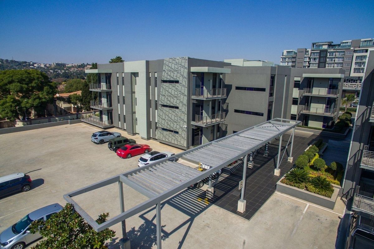 Aerial view of a modern commercial office complex featuring a multi-story building, paved parking area with covered bays, and landscaped garden sections.