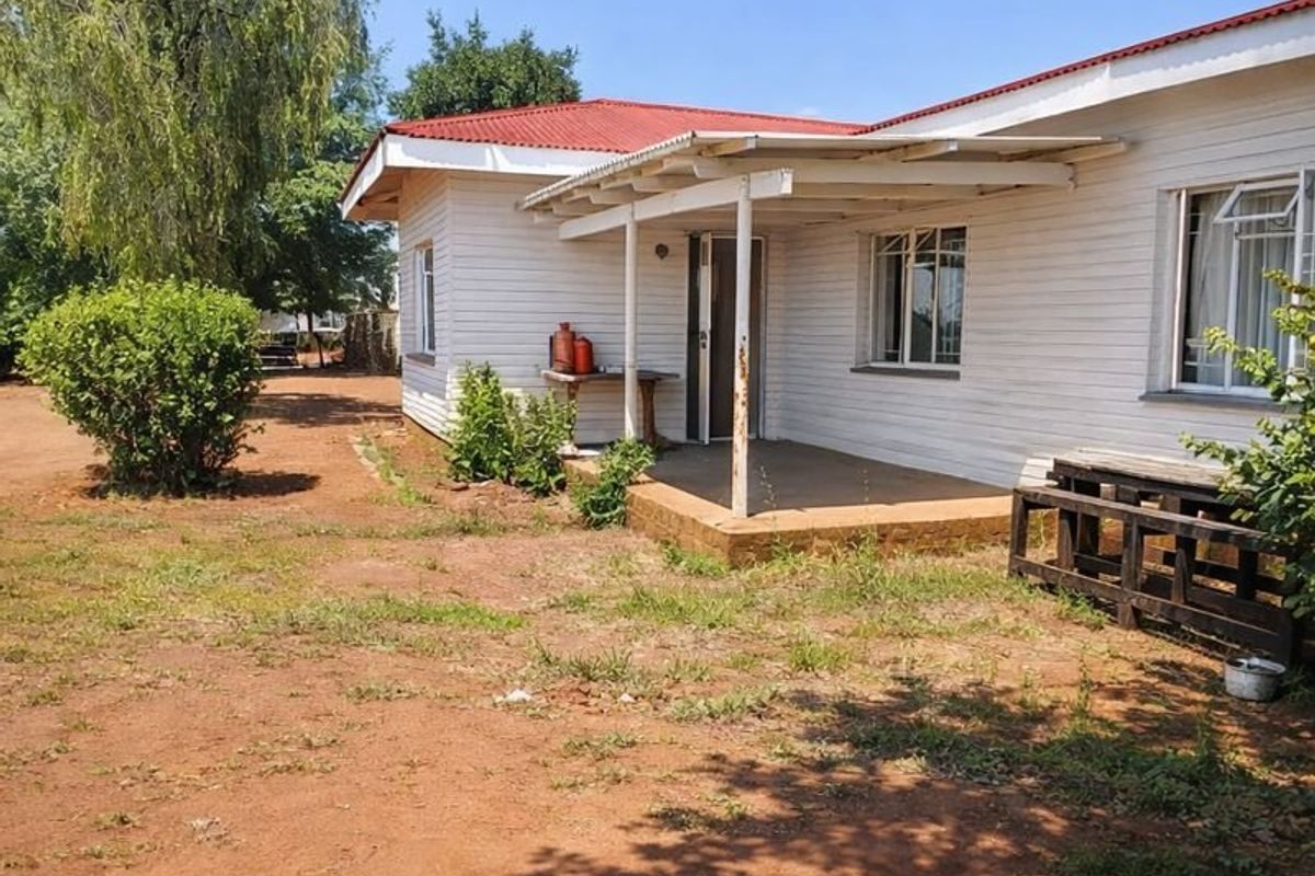 Rear view of a single-story house featuring a covered patio, corrugated metal roof, and timber siding, set on a large unpaved plot.