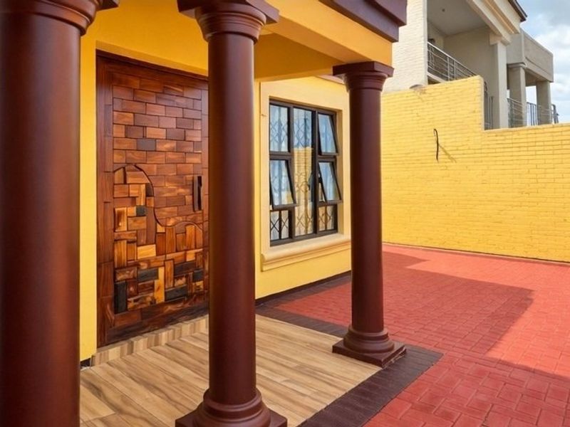 Front entrance featuring a unique decorative wooden door, structural columns, tiled porch area, and paved driveway with a yellow brick boundary wall.