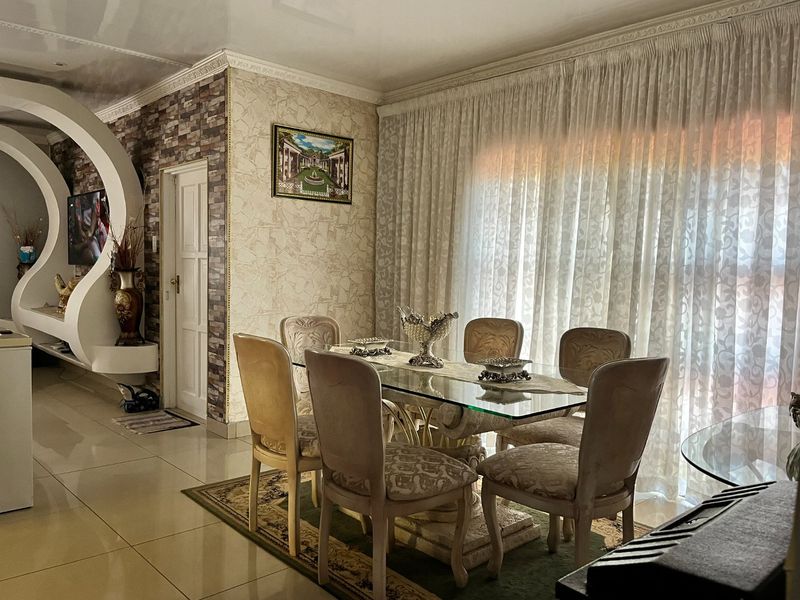 A dining room featuring tiled floors, decorative ceiling cornices, and a textured feature wall, illuminated by natural light through large curtained windows.