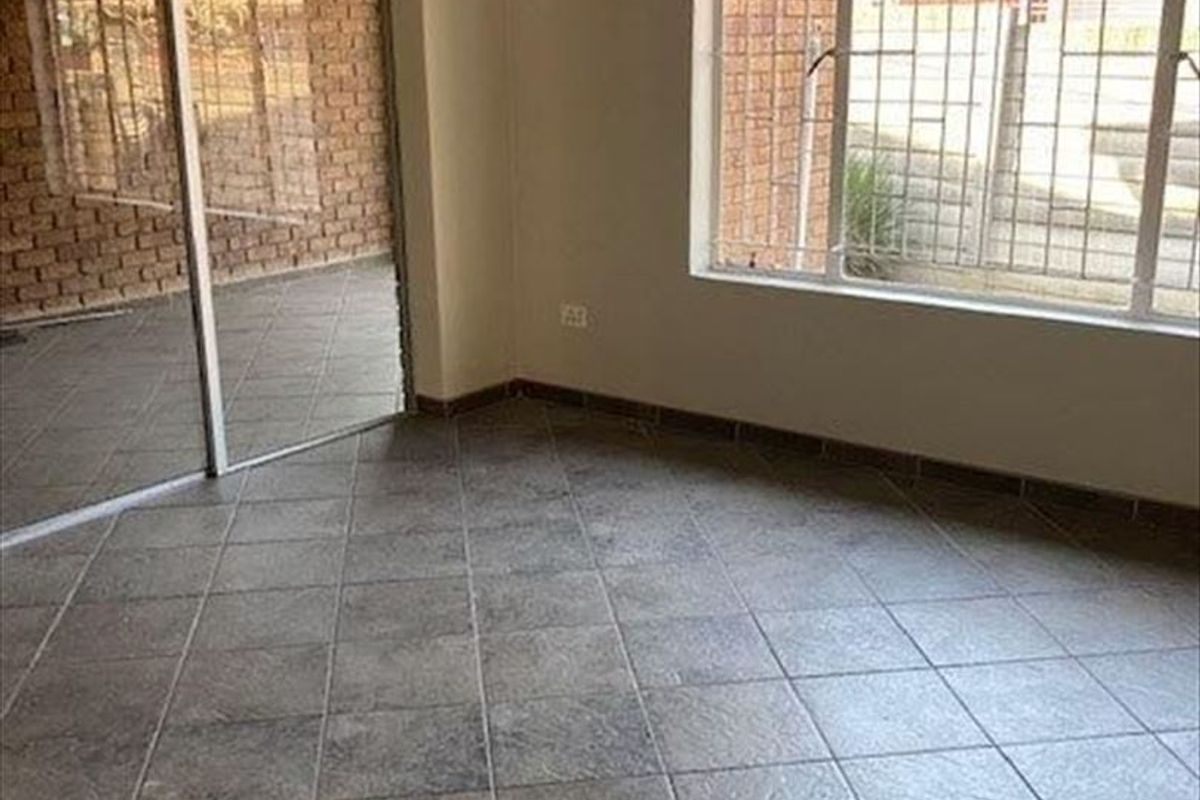 An empty living area featuring neutral tiled flooring, large windows with security bars, and a sliding glass door providing access to the exterior.