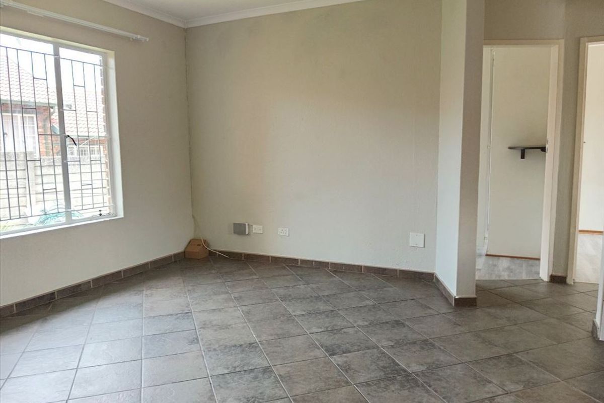 An empty living room featuring neutral tiled flooring, security bars on the window, and access to an adjacent hallway.