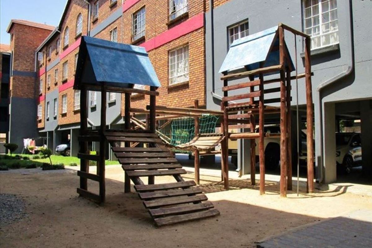 Communal outdoor area featuring a wooden playground structure situated between apartment building blocks with covered parking bays.