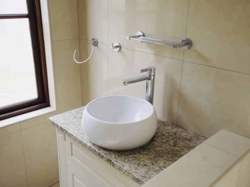 A clean bathroom vanity area featuring a modern white vessel sink, granite countertop, and chrome fixtures set against neutral wall tiles.