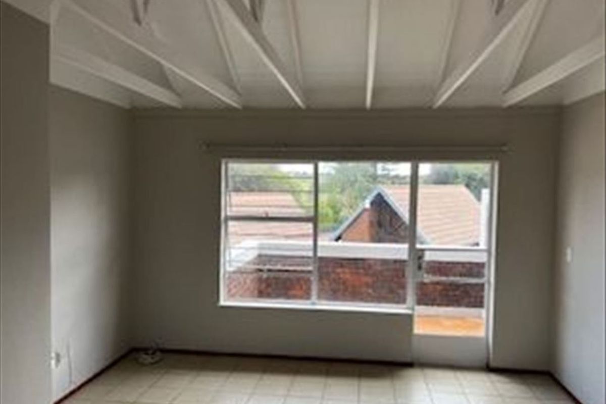 A bright, empty bedroom featuring exposed ceiling beams, tiled flooring, and a large window with access to an outdoor balcony area.