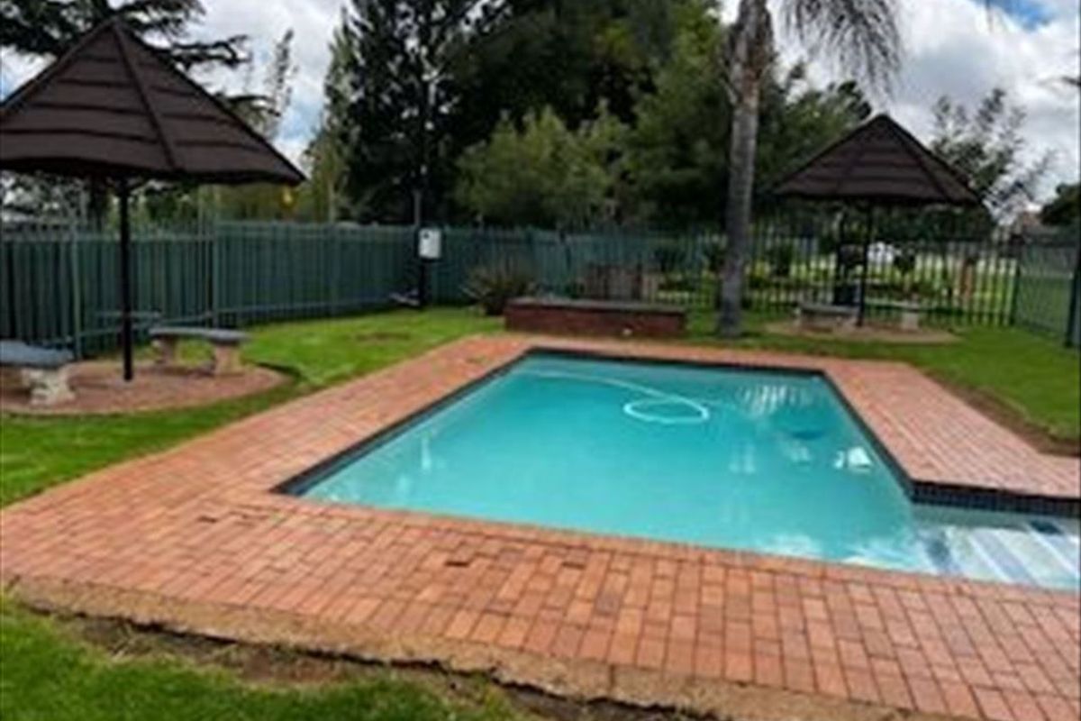 Outdoor pool area featuring a swimming pool with brick coping, fixed thatched umbrellas, stone seating, and perimeter fencing within a landscaped garden setting.