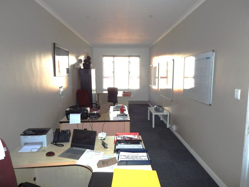 A functional office space featuring neutral walls, recessed downlighting, and carpeted flooring, providing a practical work environment with natural light from a large window.