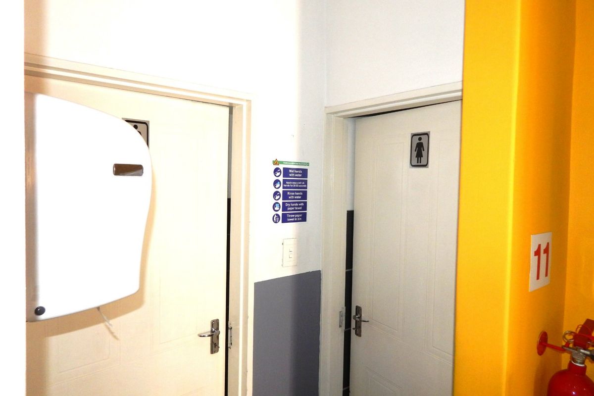 A functional industrial restroom entrance area featuring standard doors, signage, and wall-mounted safety equipment including a fire extinguisher and paper towel dispenser.